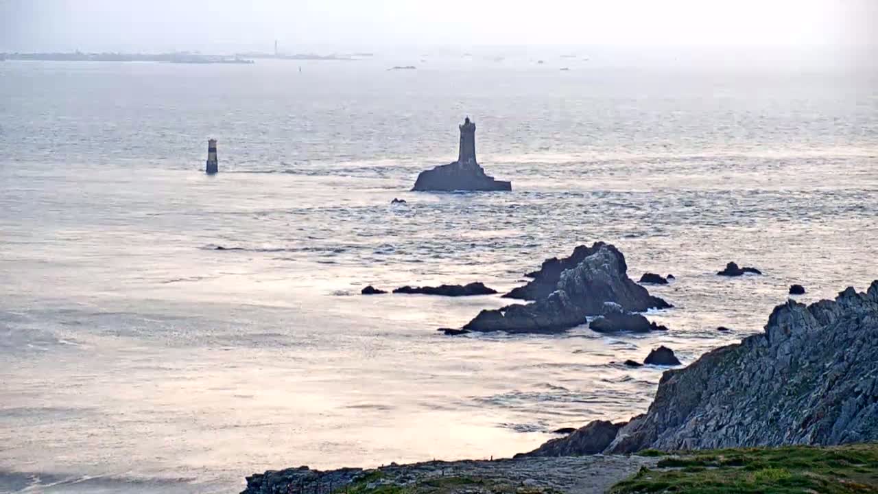 A distant lighthouse and rocky outcroppings are visible on a calm, light-grey ocean under a hazy sky.