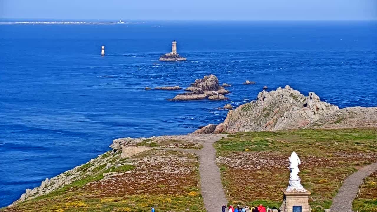 A coastal scene on a sunny day shows a rocky, grassy headland overlooking a blue ocean with several small islands and lighthouses visible in the distance.
