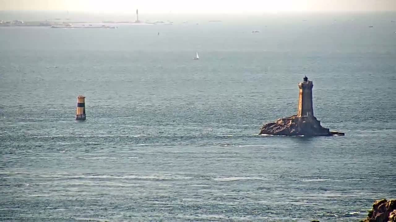 Two lighthouses stand in a calm, hazy ocean, with a sailboat visible in the distance.