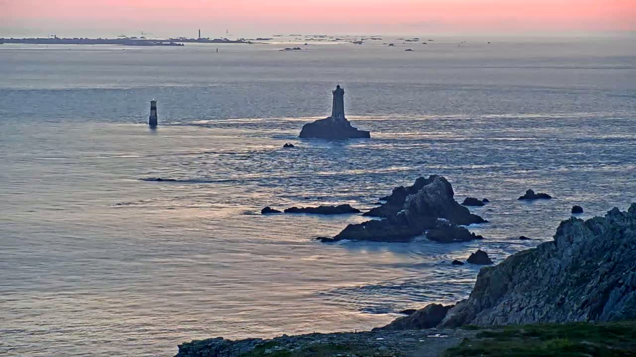 A coastal scene at sunset shows two lighthouses, one on a small island and another further away, with dark rocky outcrops along the shoreline and calm water.