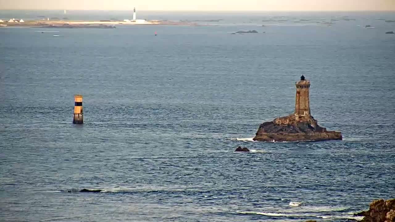 A stone lighthouse sits on a small, rocky island in a calm, blue ocean, with another smaller marker further away and a distant landmass visible on the horizon.