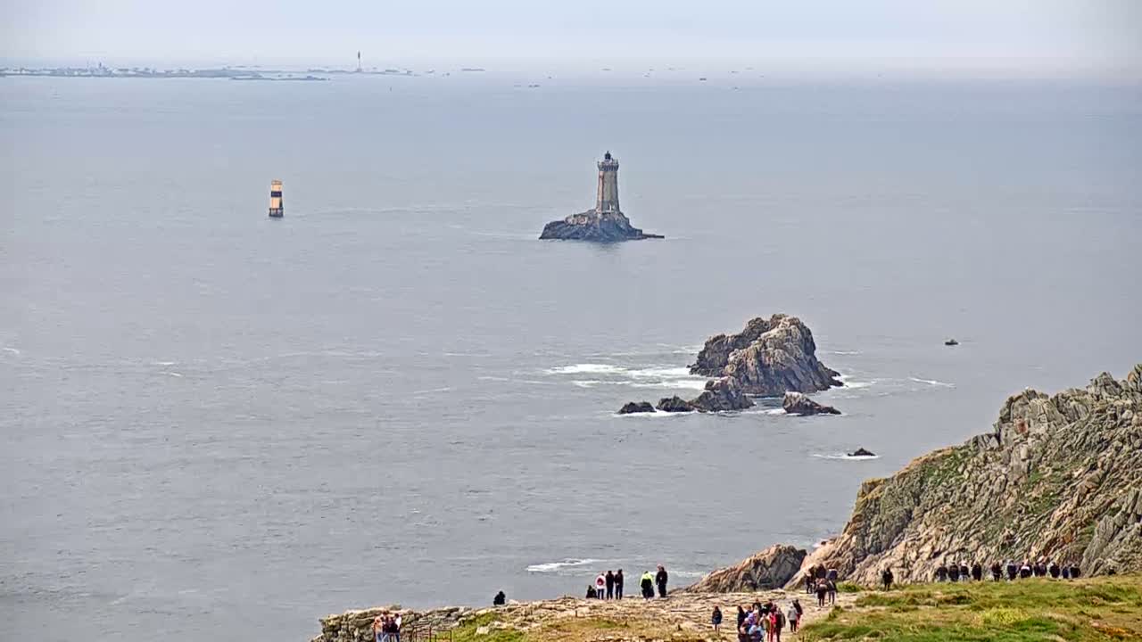 A coastal scene shows a lighthouse on a small island in the ocean, with rocky outcroppings and a group of people on a cliff overlooking the water, under hazy conditions.