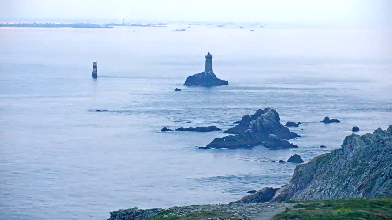 A coastal scene shows a lighthouse on a small island in a misty, calm sea with dark rocky outcrops and a distant coastline.