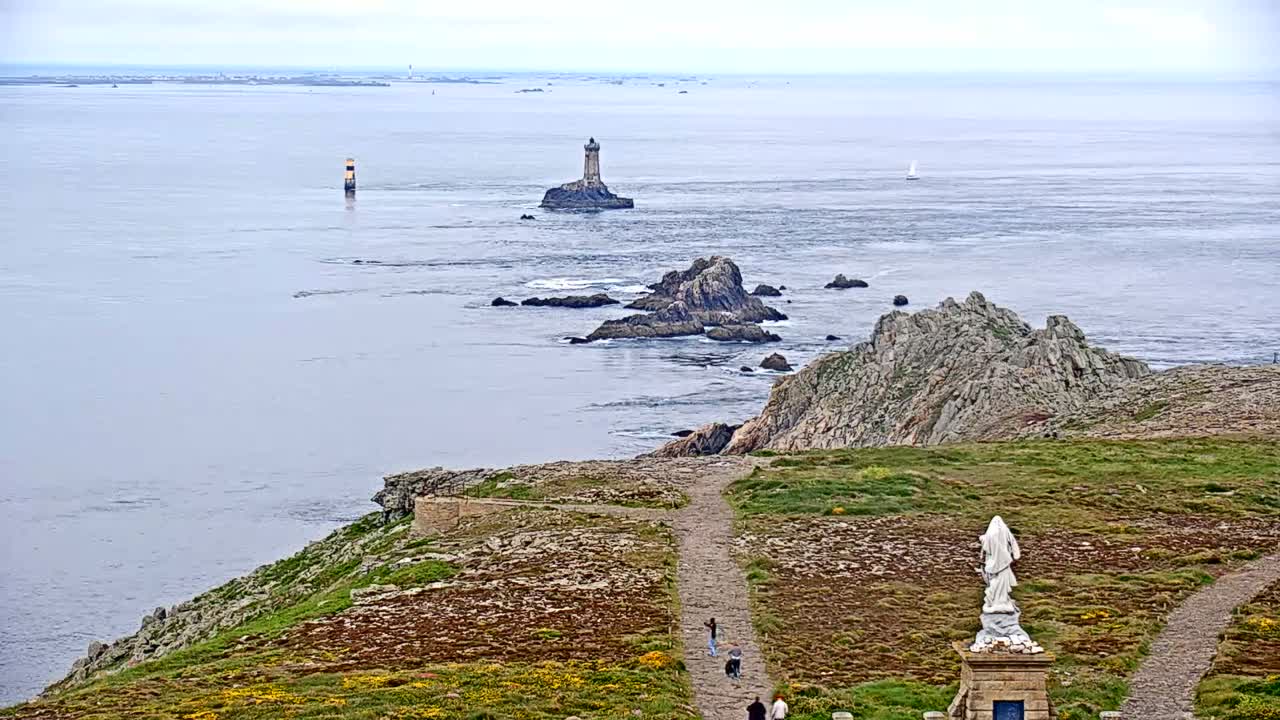 A coastal path winds through rocky terrain overlooking a body of water, featuring a distant lighthouse on a small island and a statue on the foreground, under an overcast sky.