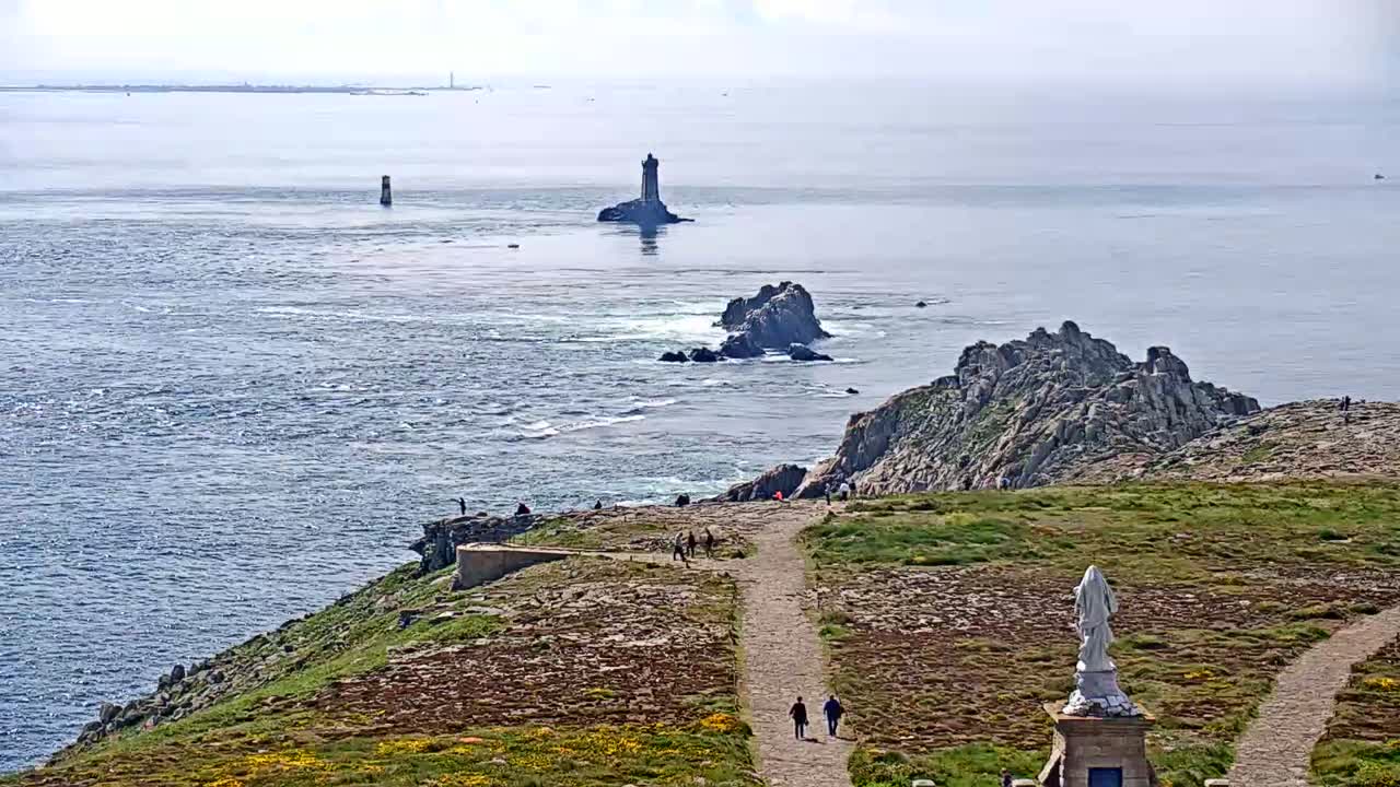 A coastal path winds down a rocky hillside toward a statue, overlooking a sea dotted with small islands and lighthouses under a hazy sky.