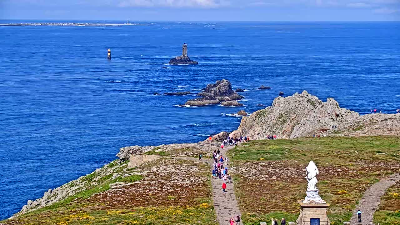A coastal path winds along a rocky cliff overlooking a vast blue ocean, where two lighthouses and several rocky outcrops are visible under a sunny sky.