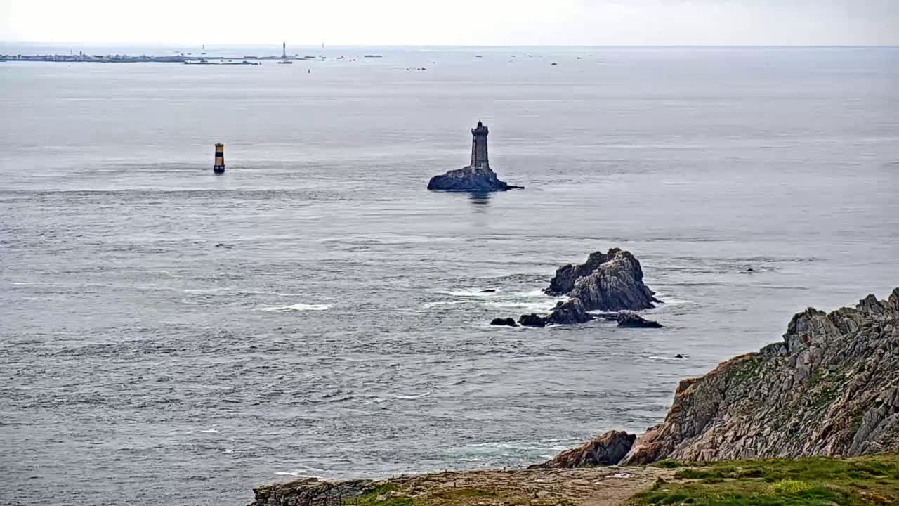 A rocky coast overlooks a calm, gray ocean with a small stone lighthouse on an island and a tall, thin marker in the distance.