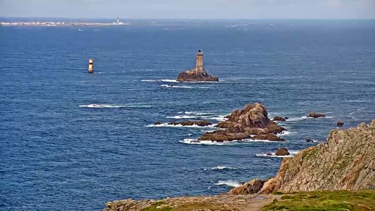 A rocky coastline with a lighthouse on a small island in the distance, under a partly cloudy sky, with waves crashing against the rocks.