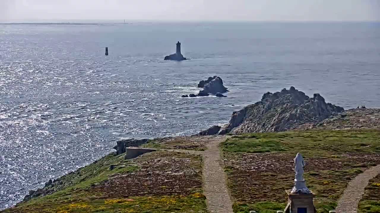 A statue stands on a grassy coastal path overlooking a rocky shoreline and a calm, sunlit ocean with several small islands and lighthouses in the distance.