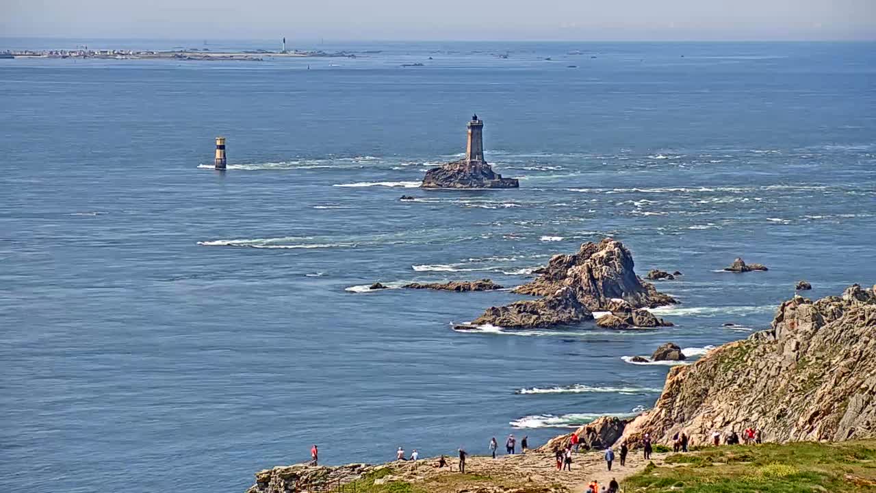 A coastal scene on a sunny day shows a lighthouse on a small island surrounded by rocky outcrops and the ocean, with people gathered on a cliff overlooking the water.