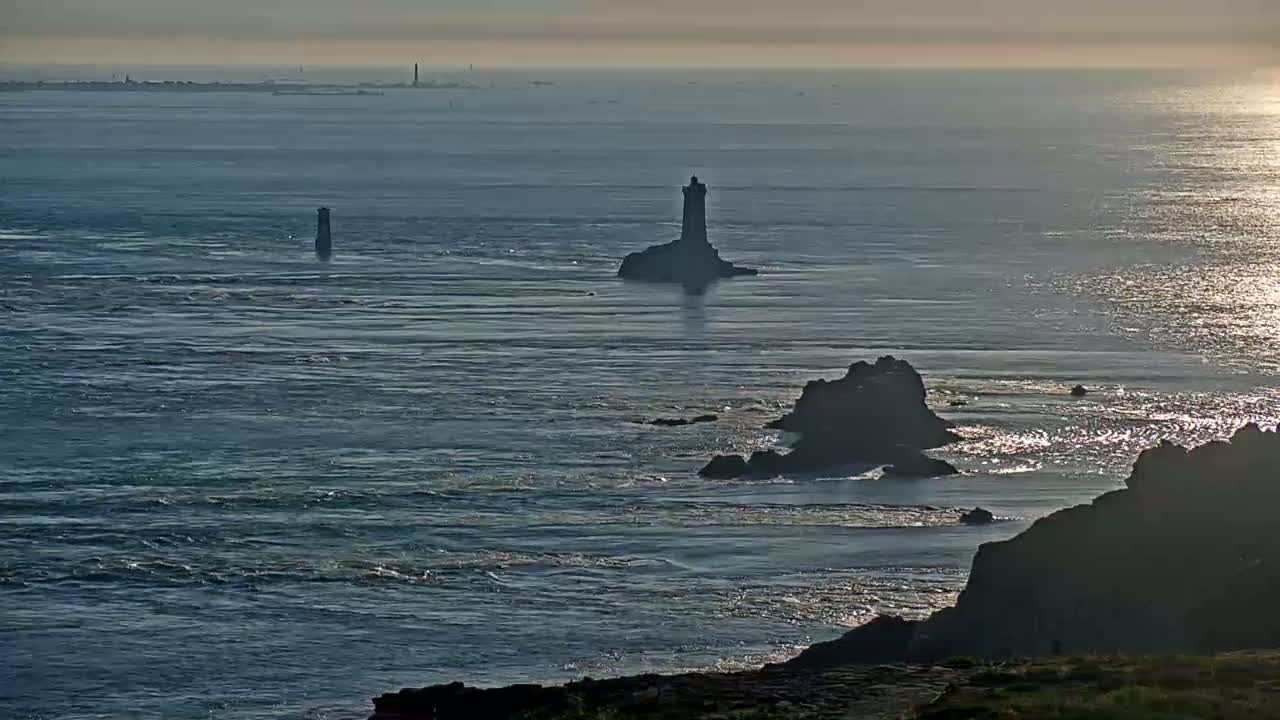 A lighthouse sits on a small island in a choppy sea, with other rocky outcrops and a distant coastline visible under a mostly cloudy sky.