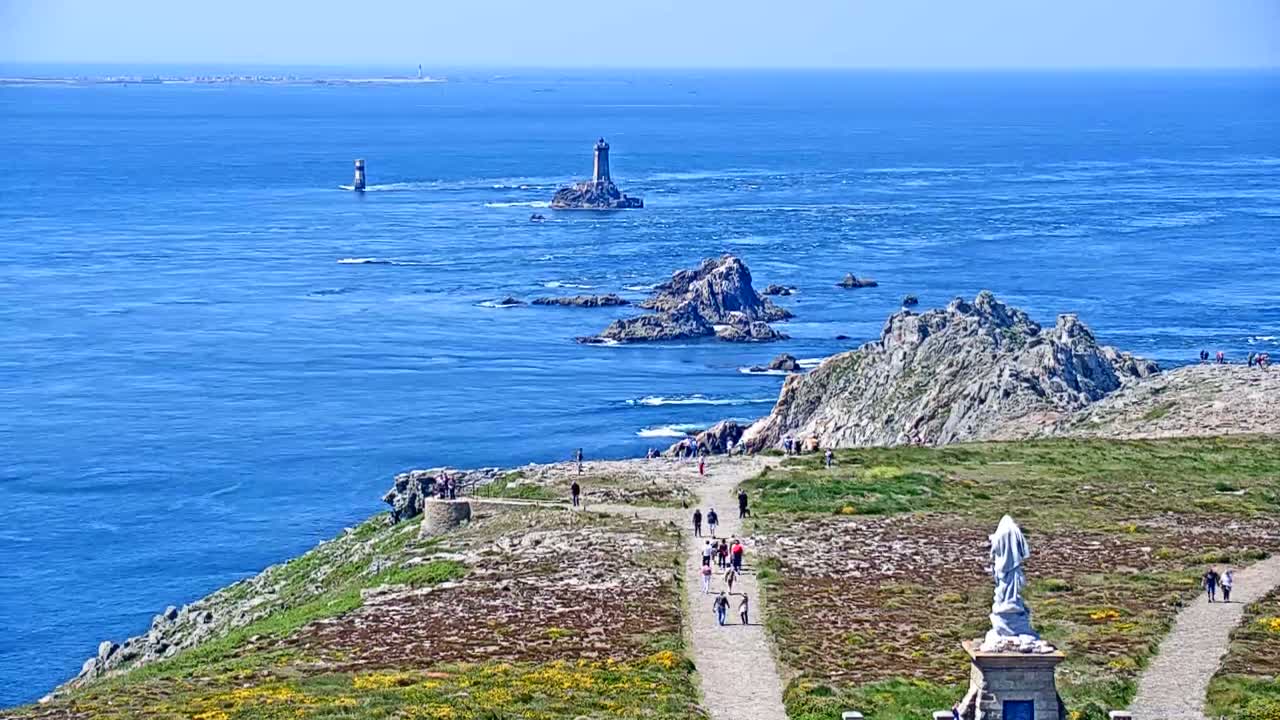 A coastal scene on a sunny day shows a path winding along a rocky cliff overlooking a vast expanse of blue ocean dotted with islands and lighthouses.