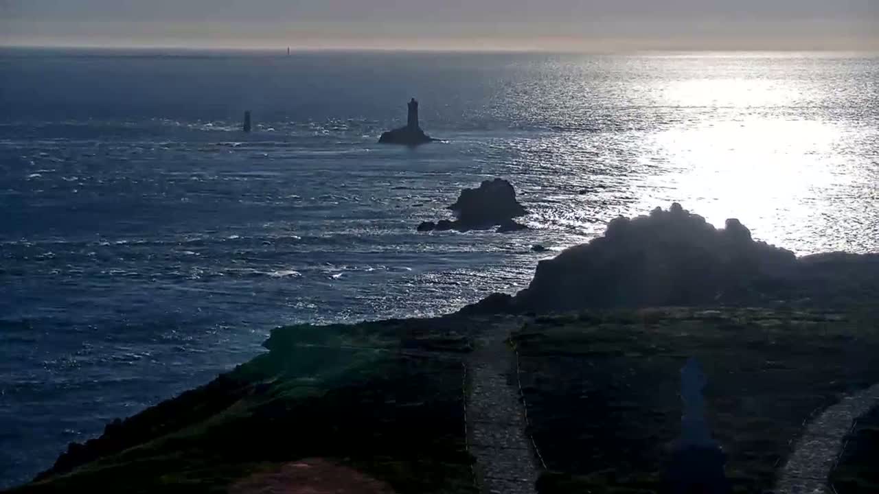 A rocky coastline with several small islands and lighthouses is visible under a bright, sunny sky, with a somewhat choppy sea.