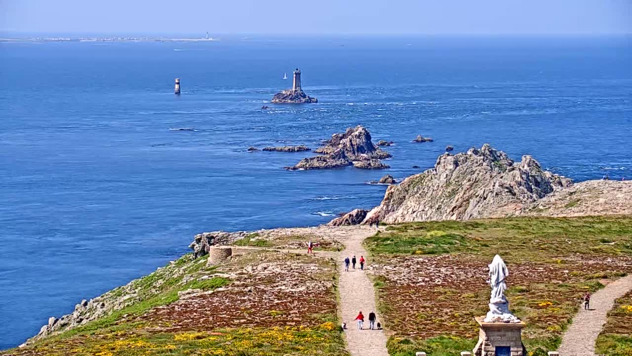 A coastal scene on a sunny day shows a rocky shoreline with a statue and several people walking along a path overlooking the ocean, where several islands and lighthouses are visible in the distance.