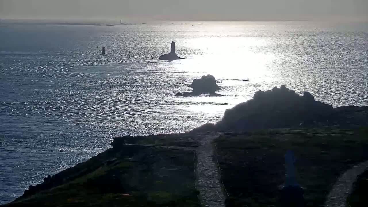 A rocky coastline with several lighthouses visible in the distance under a bright, sunny sky, with a gently rippling sea reflecting sunlight.