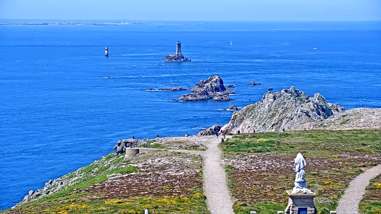 A sunny day reveals a coastal scene with a lighthouse in the distance, rocky outcrops, a statue, and winding paths on a grassy cliff overlooking a vibrant blue ocean.