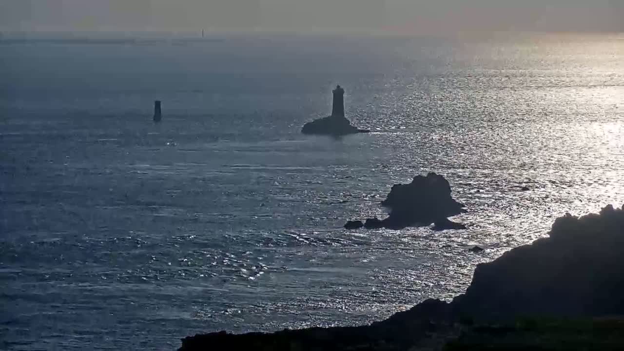 A dark, rocky coastline with a lighthouse and smaller rock formations in a glistening sea under a hazy sky.