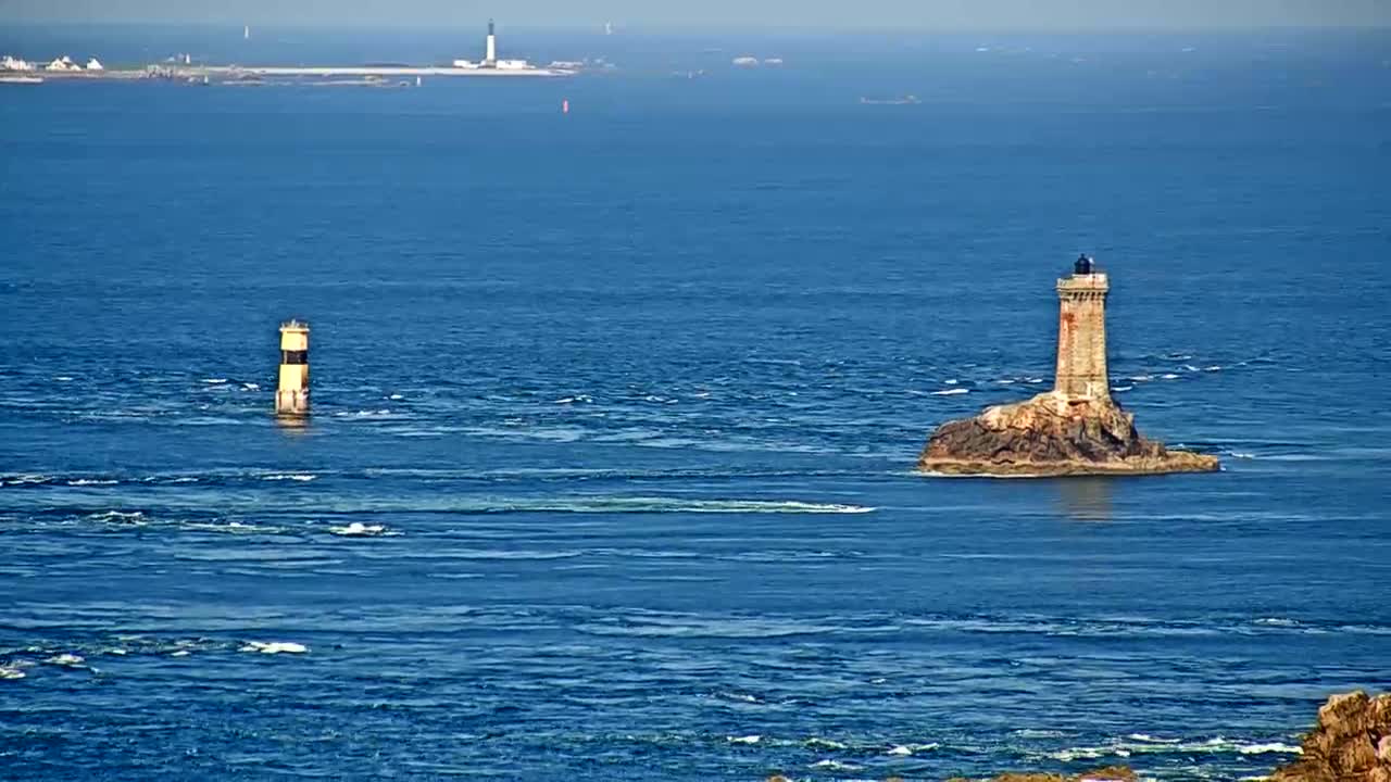 A distant island with a lighthouse is visible on a calm, blue ocean, with another smaller, cylindrical lighthouse in the foreground.