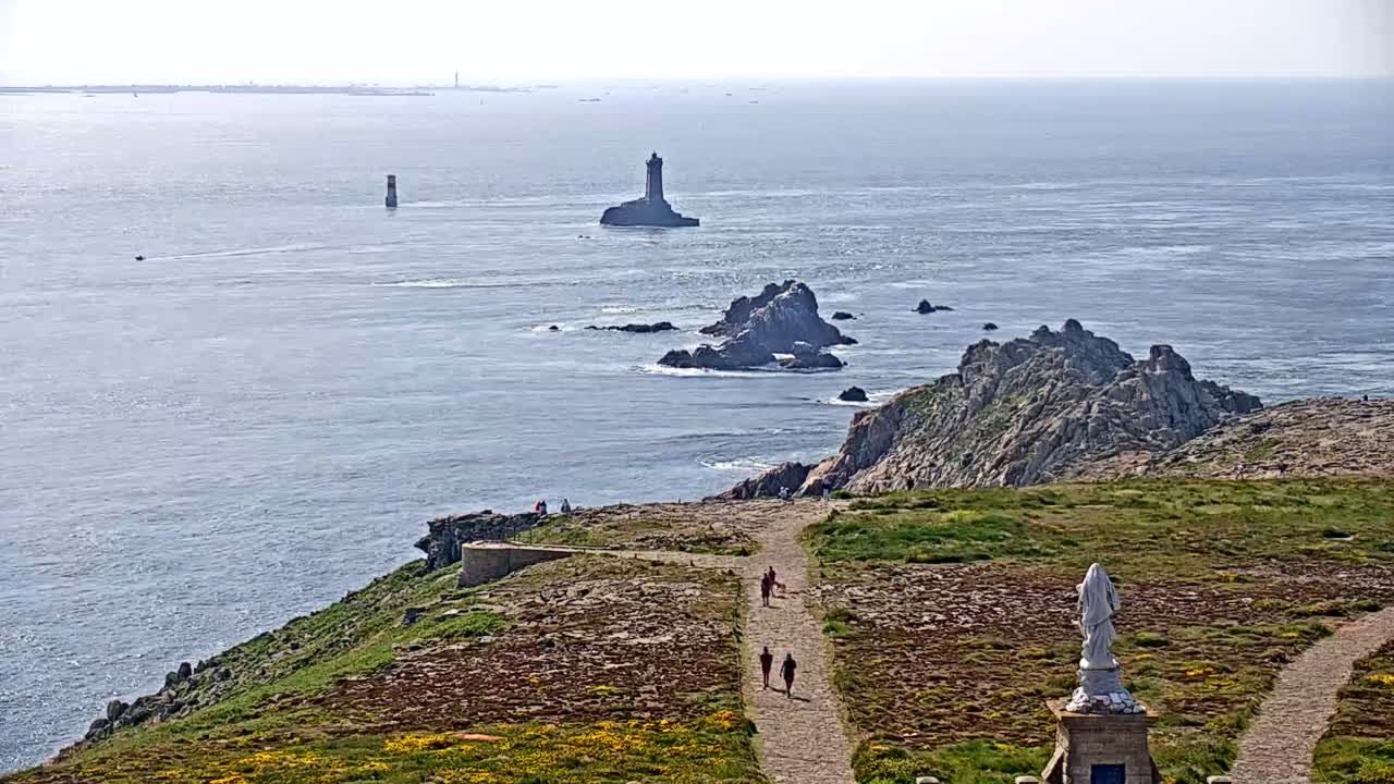 A coastal scene on a sunny day shows a rocky, grassy cliff overlooking a body of water with several small islands, one featuring a lighthouse, and a statue visible on the cliff.