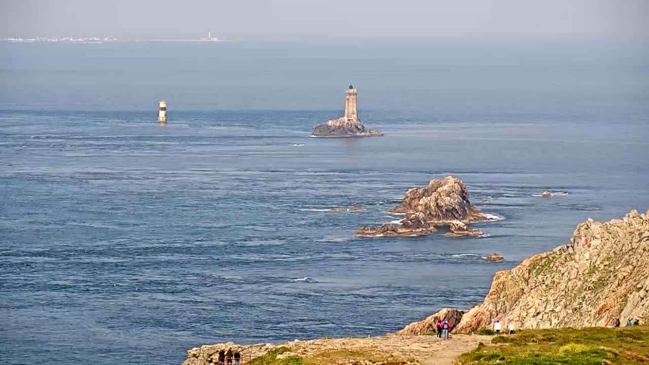 A coastal scene on a clear day shows a lighthouse on a small island, other rocky outcrops, and a group of people standing on a cliff overlooking the ocean.