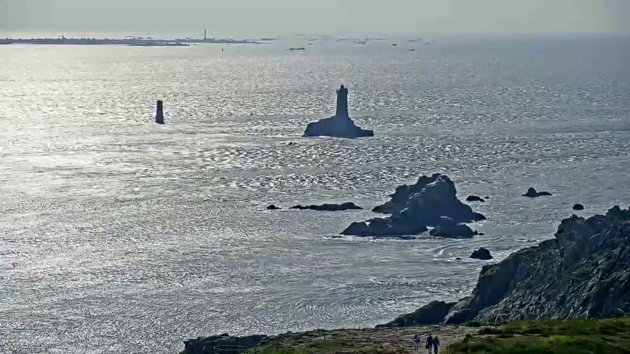 A coastal view shows a lighthouse on a small island surrounded by dark, rocky outcrops and a calm, sparkling sea under a hazy sky.