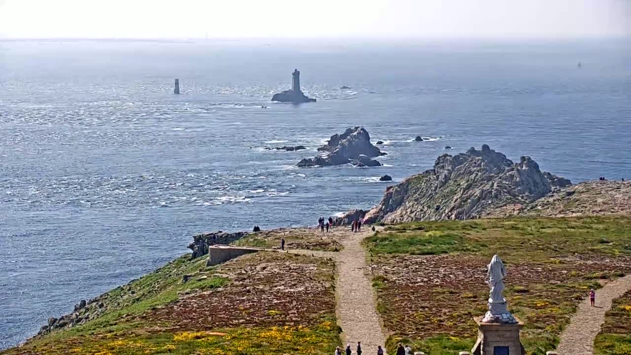A coastal scene on a sunny day shows a rocky outcrop with a statue and a path, overlooking a body of water with several small islands and lighthouses in the distance.