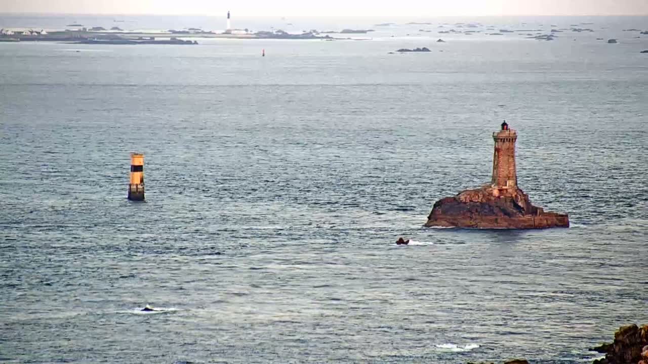 A stone lighthouse sits on a small, rocky island in a calm sea, with a smaller, orange and black navigational marker visible in the distance.