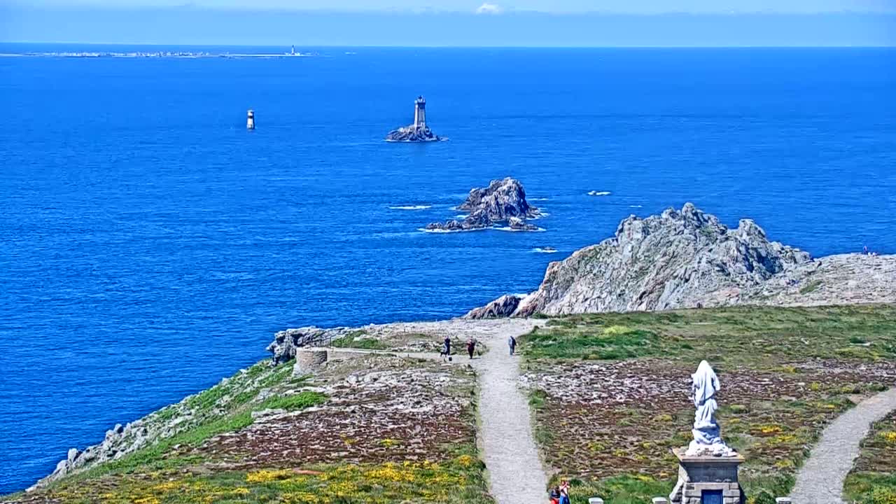 A coastal path winds through low-lying vegetation toward a statue overlooking a blue ocean dotted with rocky outcrops and distant lighthouses under a sunny sky.