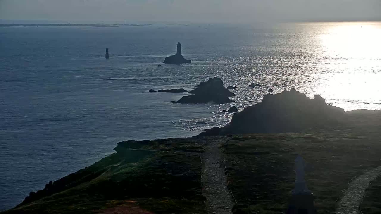 A rocky coastline with a lighthouse and other structures is visible against a bright, sunlit ocean under a mostly cloudy sky.