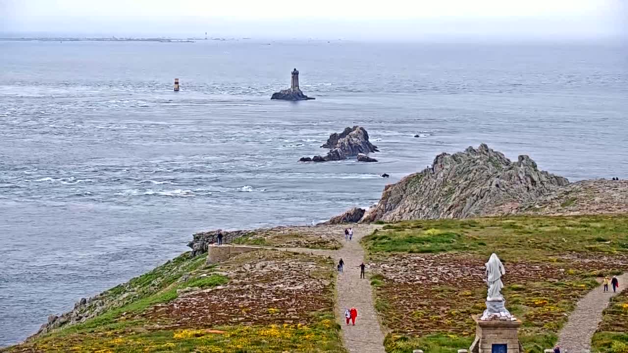 A coastal scene on an overcast day shows a rocky shoreline with a path leading to a statue, with a distant lighthouse and several small islands visible across the water.