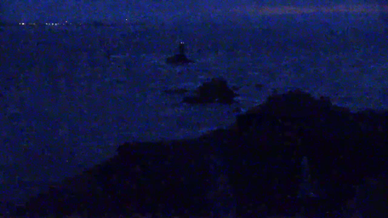 A dark, nighttime scene shows a lighthouse and rocky outcrops in a choppy sea, with a distant city skyline visible on the horizon.