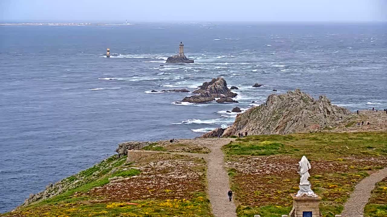 A rocky coastal path overlooks a choppy ocean with several lighthouses visible in the distance, and a statue stands near the path.