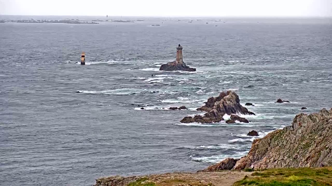 A rocky coastline with a lighthouse on a small island and a sea marker in the distance, under overcast skies and slightly choppy waters.