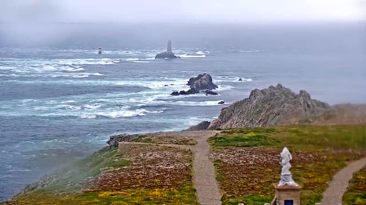 A foggy coastal scene shows a rocky shoreline with a statue, a path, and a distant lighthouse surrounded by a choppy sea.