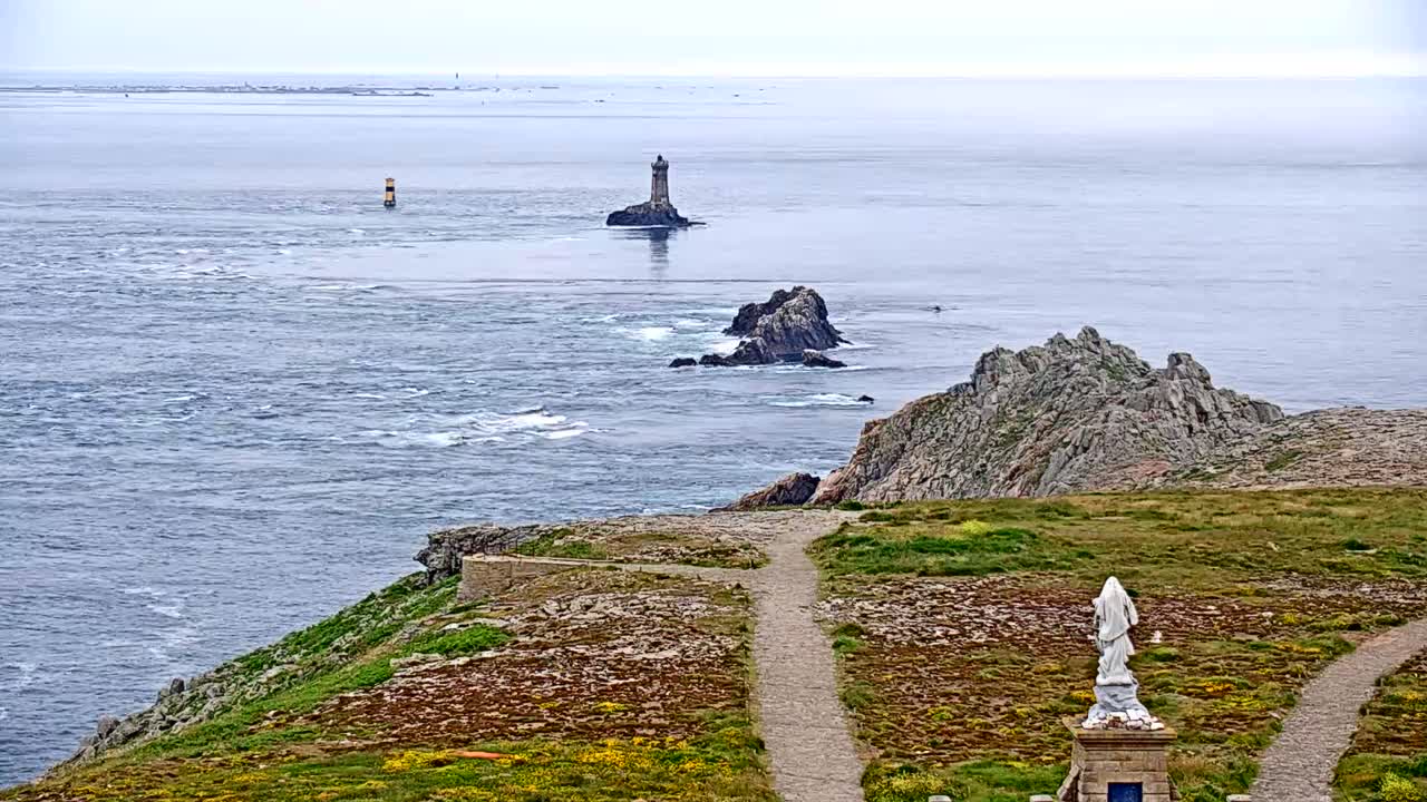 A coastal scene on an overcast day shows a rocky shoreline with a statue, a path, and a distant lighthouse on a small island.
