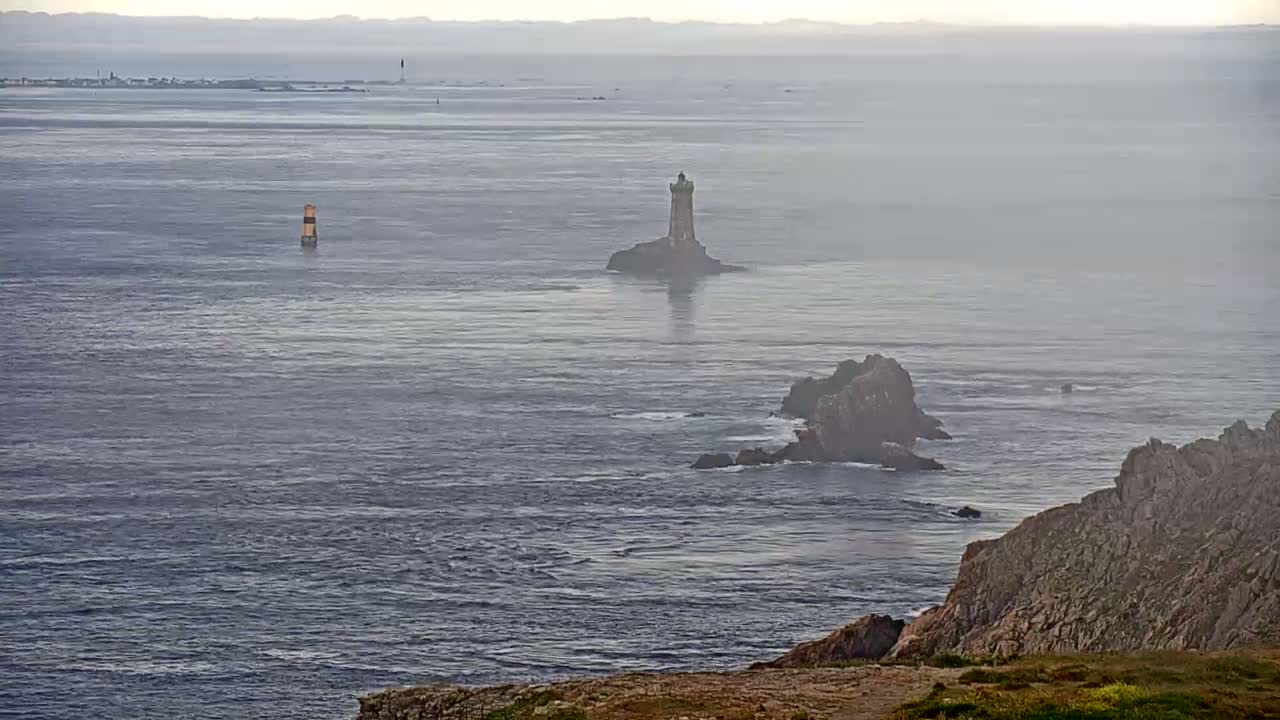 A hazy coastal scene shows a lighthouse on a small island, rocky outcrops, and a distant shoreline in a calm sea.
