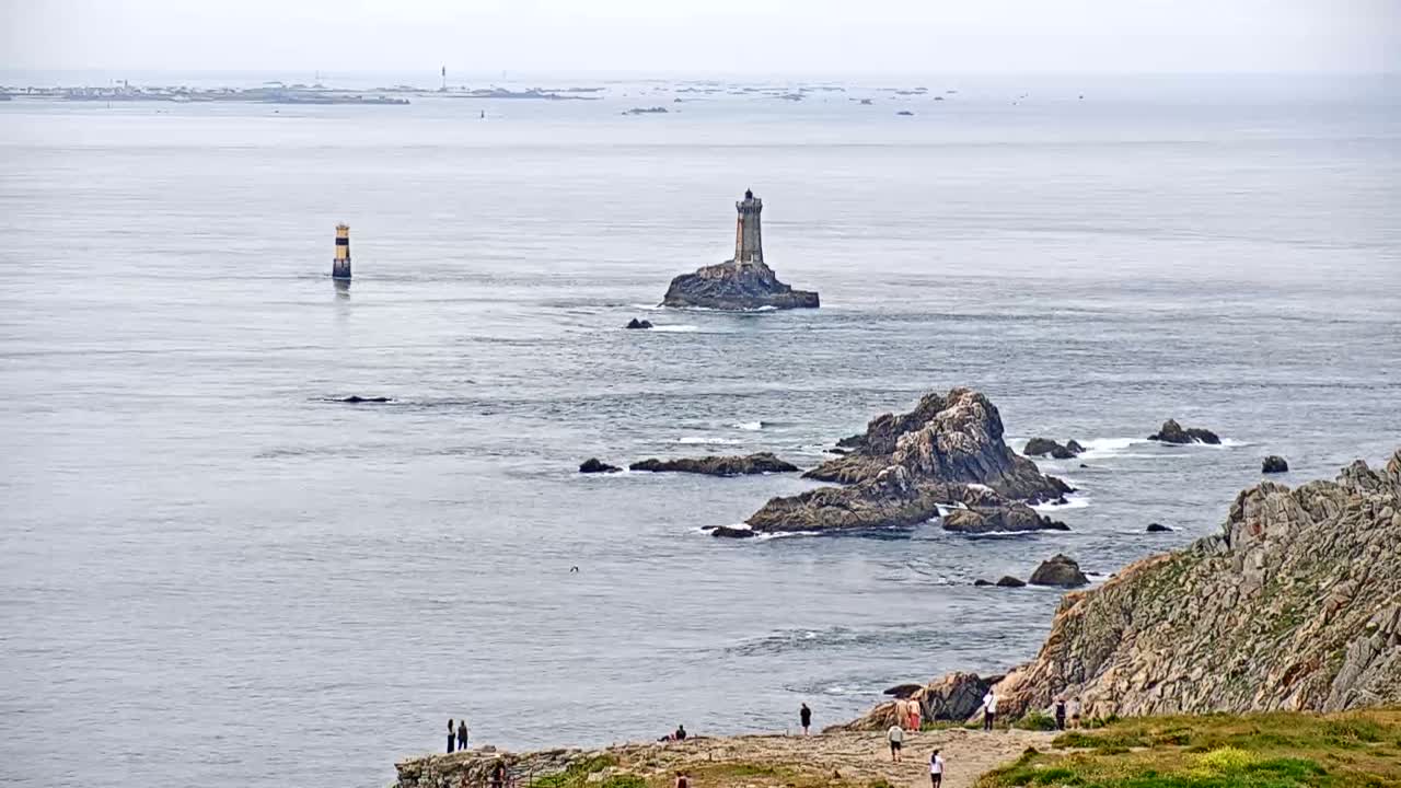A coastal scene on an overcast day shows a lighthouse on a small island, rocky outcrops, a distant coastline, and several people standing on a cliff overlooking the ocean.