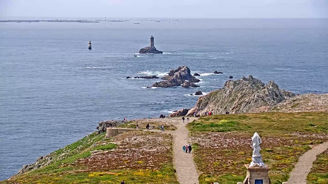 A coastal scene on a partly sunny day shows a rocky, grassy outcrop with a statue and walking paths overlooking a wide expanse of ocean with two lighthouses and distant islands visible.