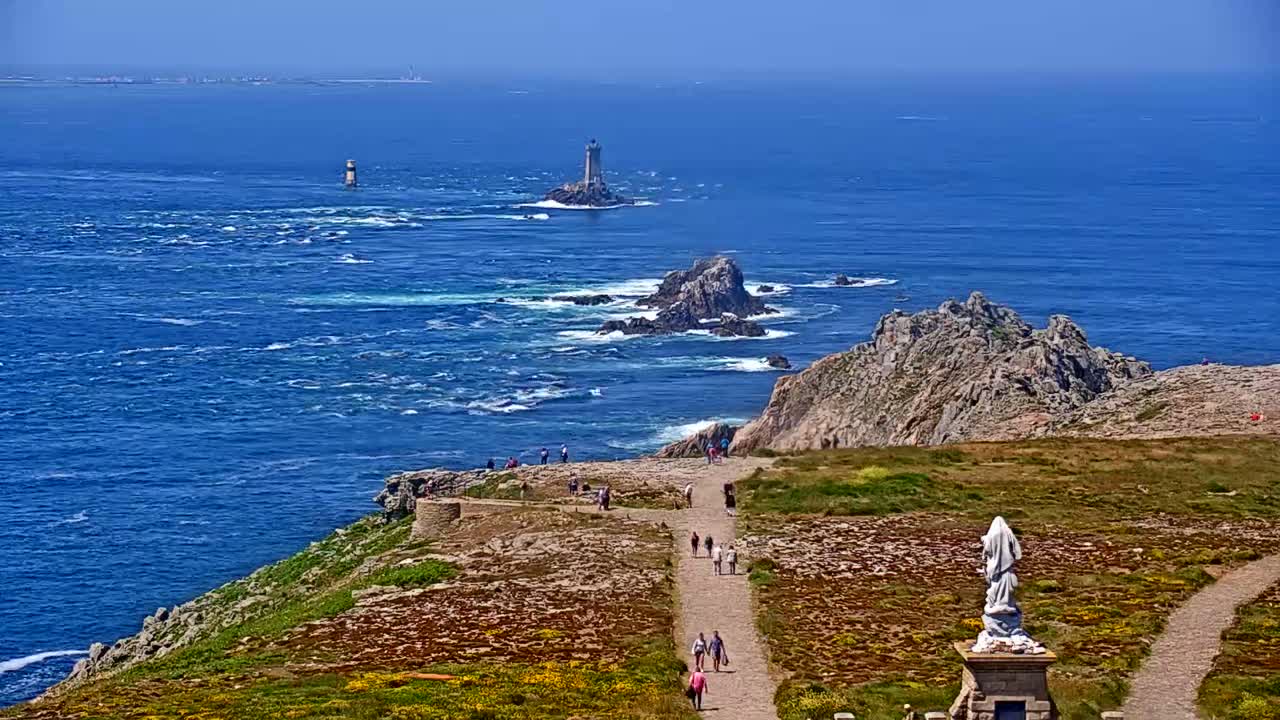 A coastal path winds through grassy terrain toward a statue overlooking a blue ocean with rocky outcrops and distant lighthouses under a clear sky.