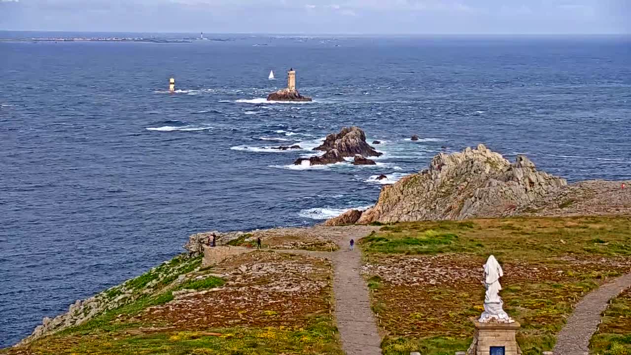 A coastal scene on a partly cloudy day shows a rocky, grassy headland with a statue, paths, and a distant view of lighthouses and islands across a choppy sea.