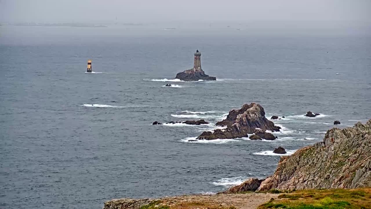A lighthouse sits on a small island in a choppy sea under an overcast sky, with rocky outcroppings visible in the foreground and another marker in the distance.