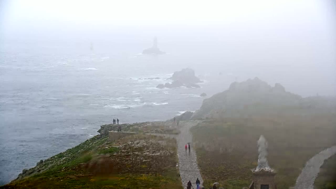 A foggy coastal scene shows a rocky path winding down a cliff toward the ocean, where several people are walking, with a distant island and lighthouse barely visible through the mist.