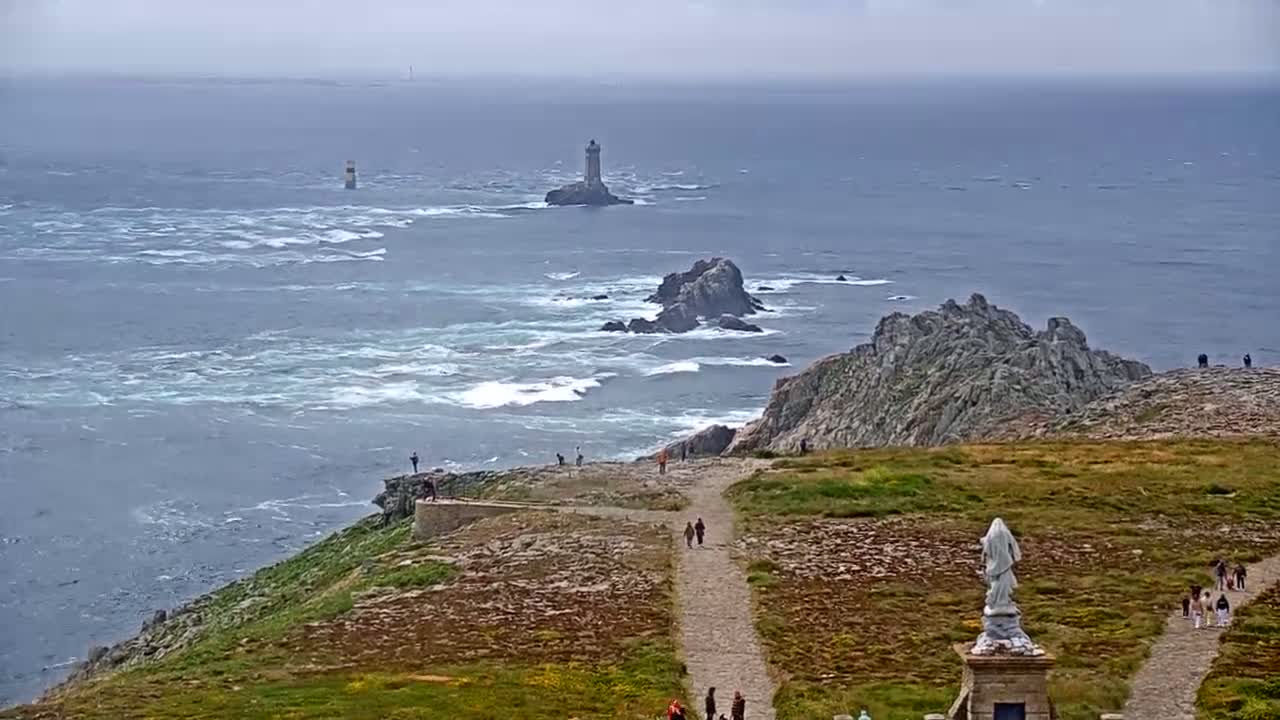 A rocky, grassy coastal area with a statue overlooks a choppy sea where two lighthouses are visible in the distance under an overcast sky.