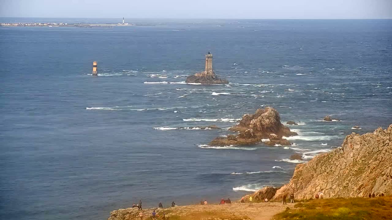 A rocky coastline with a lighthouse on a small island in a choppy sea, with a distant landmass visible under a mostly clear sky.