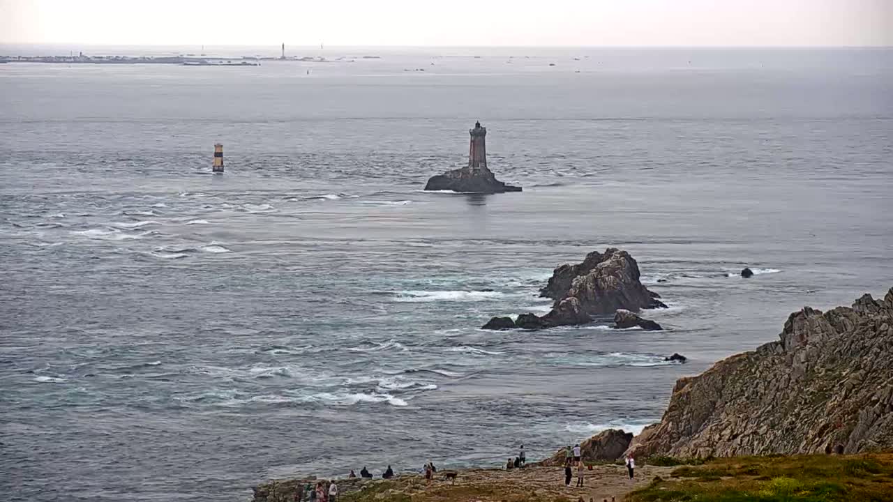 A lighthouse sits on a small island in a choppy sea, with rocky coastlines and a small group of people visible on land in the foreground, under an overcast sky.