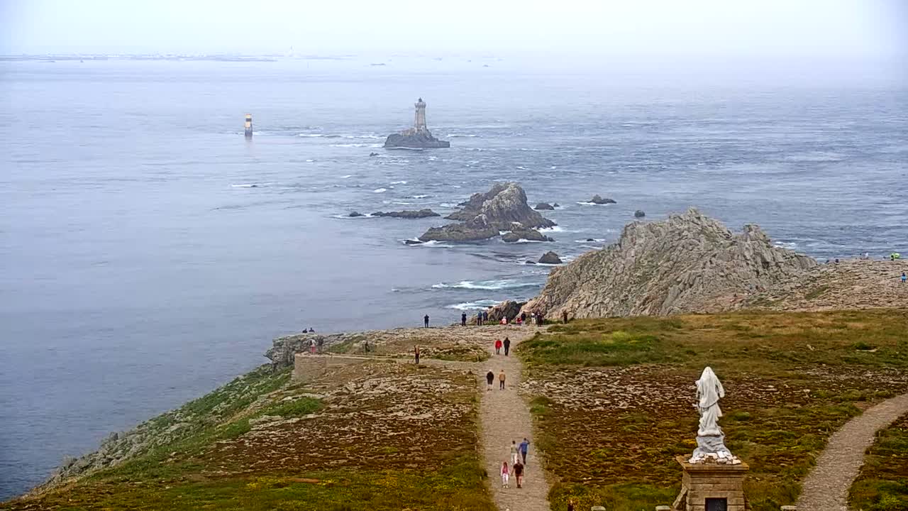 A coastal scene on a foggy day shows a rocky outcrop with a statue and several people walking on a path, overlooking a body of water with a distant lighthouse.