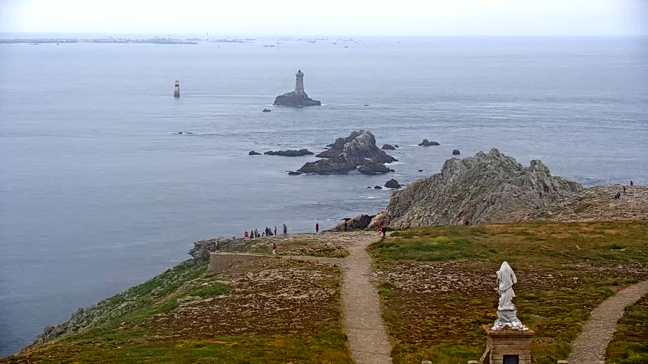 A rocky coastal scene on an overcast day shows two lighthouses in the distance, with a statue and a few people visible on a grassy path overlooking the ocean.