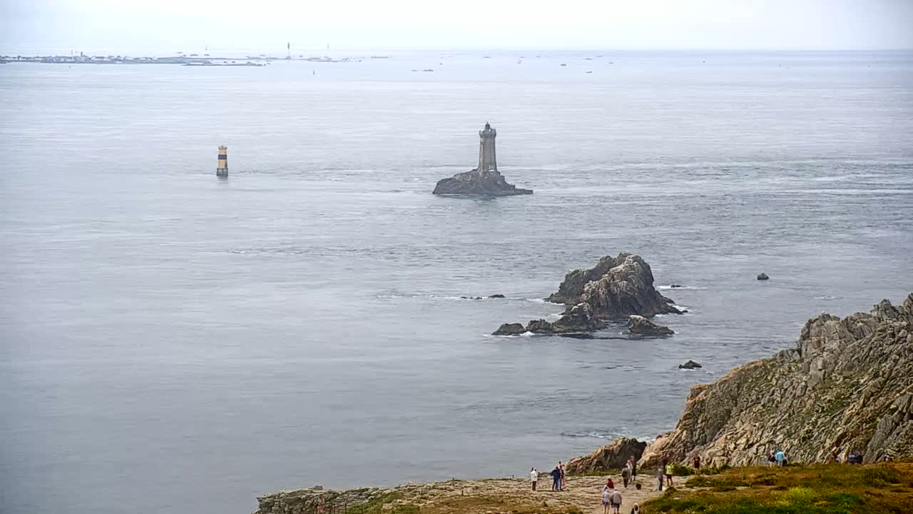 A lighthouse sits on a small island in a calm, gray sea, with rocky outcrops and a group of people on a cliff in the foreground.