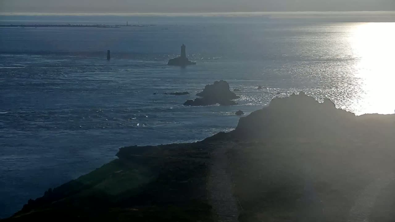 A hazy, sunlit ocean stretches to a distant horizon, featuring a lighthouse on a small island and dark rocky coastline in the foreground.