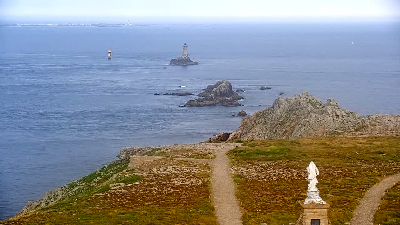 A coastal scene on a hazy day shows a statue on a grassy cliff overlooking the ocean with rocky outcrops and distant lighthouses.